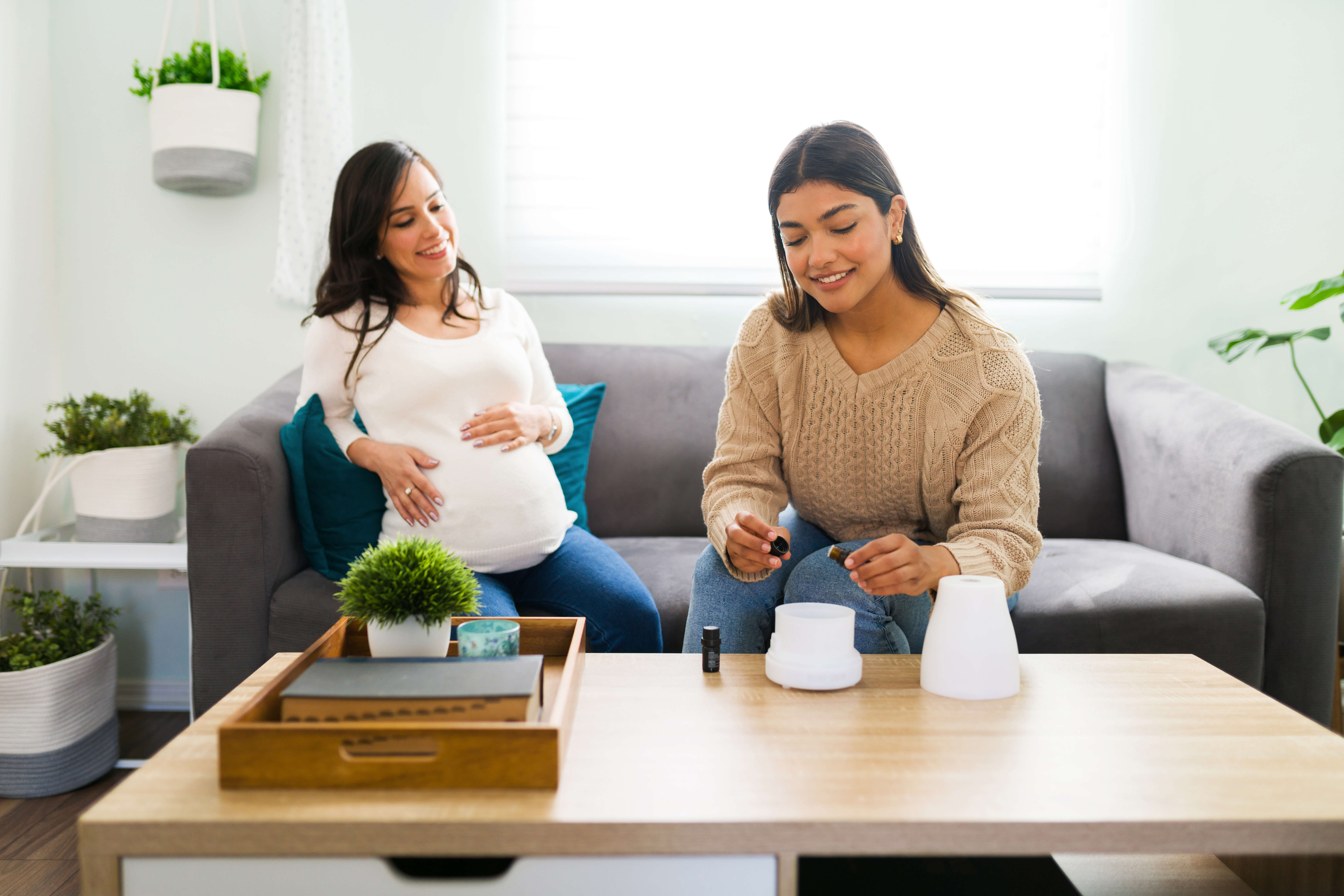 Smiling midwife giving a stressed expectant mother essential oils Happy hispanic doula putting essential oils with lavender in a diffuser during a home visit to help a caucasian pregnant woman to rest better and relax